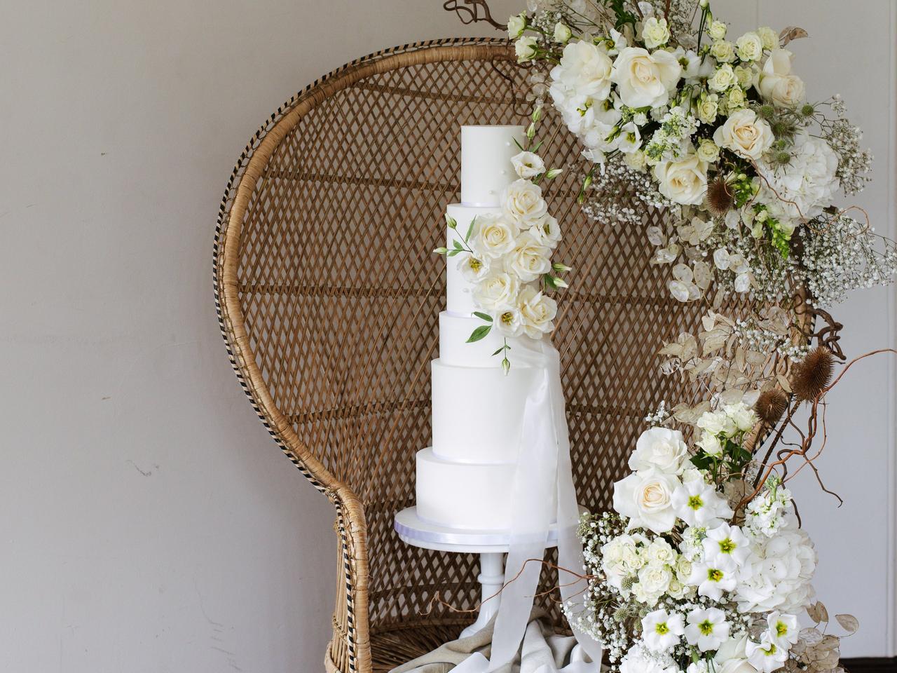 A tall white iced wedding cake with no detail except some white sugar flowers set against the backdrop of a wicker chair draped in white flowers.