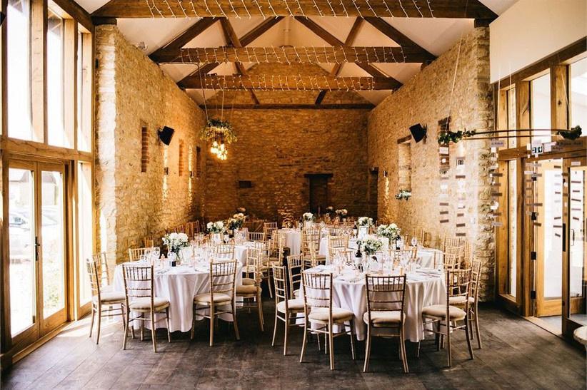 Rustic room laid for wedding meal with round white tables, Chiavari chairs, exposed brick, exposed wooden beams, fairy lights and floor-to-ceiling windows