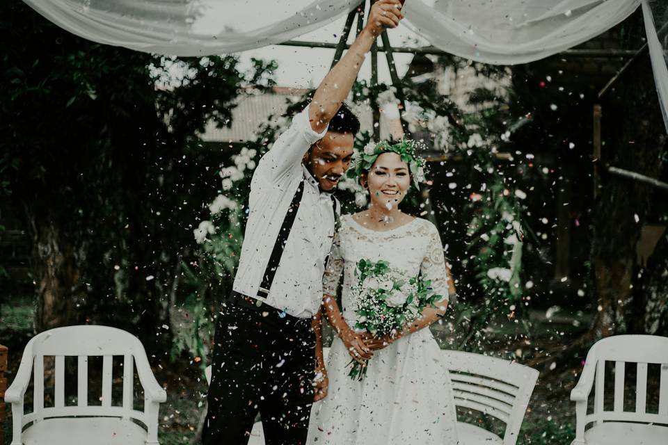 A groom holding a confetti cannon in the air as confetti cascades down over him and his bride