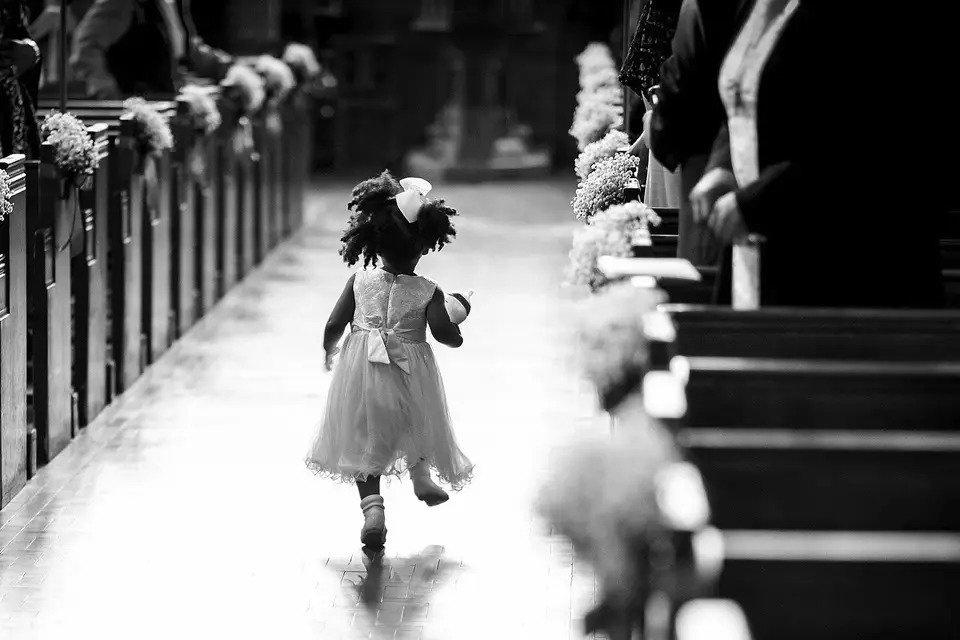 A black and white photo taken from behind of a little flower girl skipping down the aisle