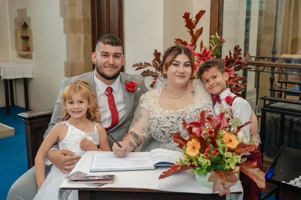 Bride, groom and two children smile at the camera while signing the register surrounded by flowers