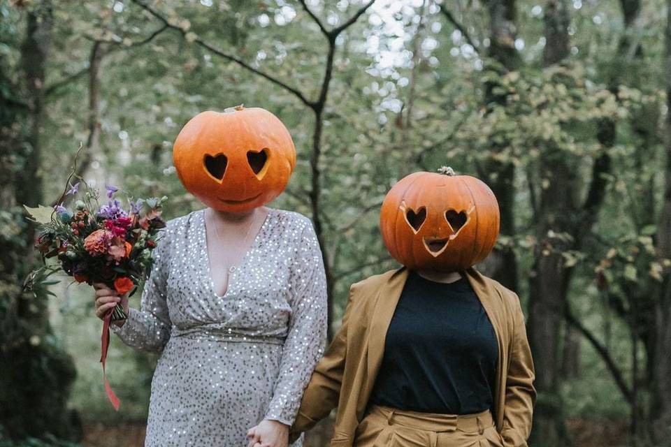 A couple wearing pumpkins with heart-shaped eyes on their heads. One is wearing a sparkly dress and holding a bouquet and the other is wearing a brown suit. They are in an autumnal forest