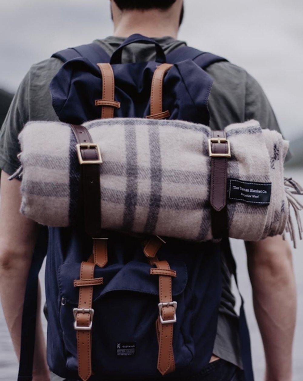 Picture of man from behind wearing a navy backpack with a neutral tartan blanket rolled at the top