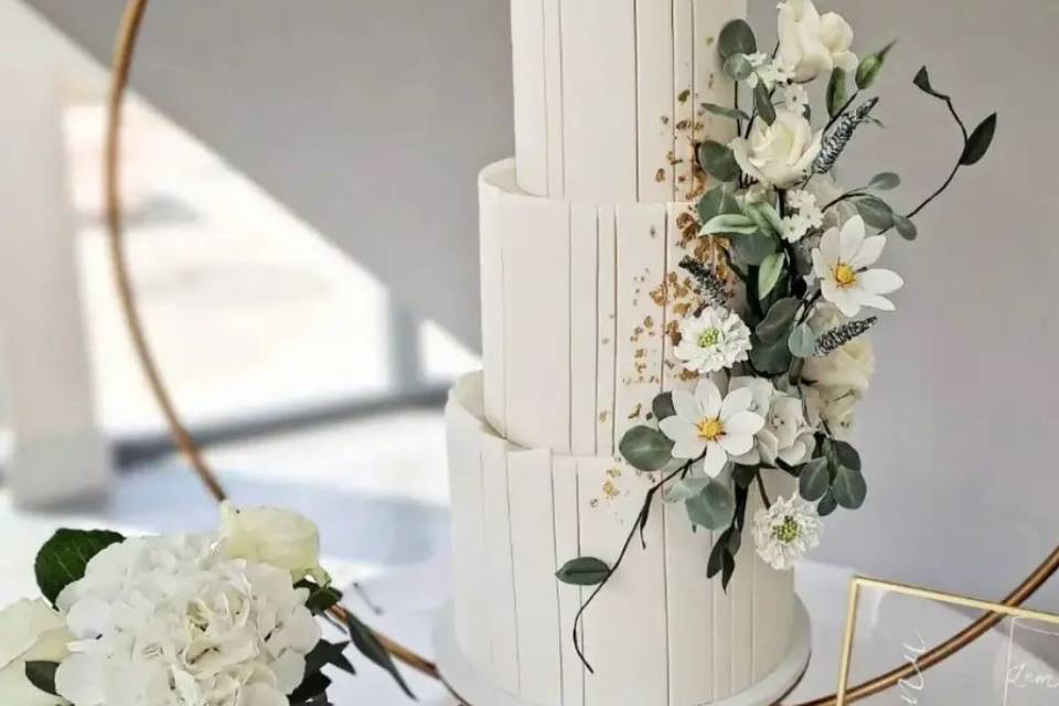 white four tier wedding cake sat on a gold hoop stand with white fondant panels for decoration as well as white and green flowers