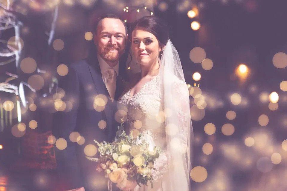 Photo of a bride and groom looking directly at the camera, with the shot edited to enhance the light flares from fairy lights