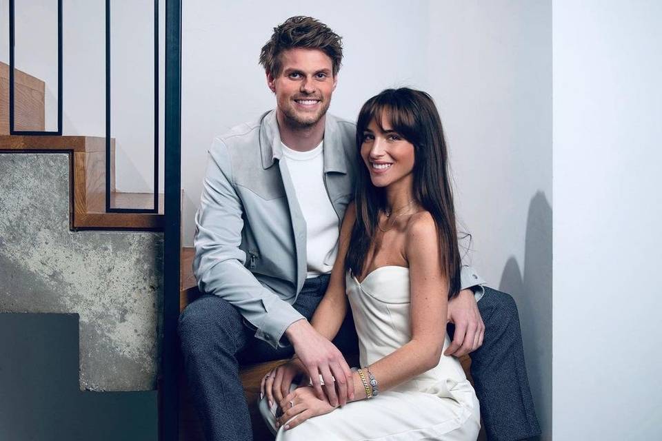 Maeva wearing a white strapless jumpsuit sat between James's legs on a staircase