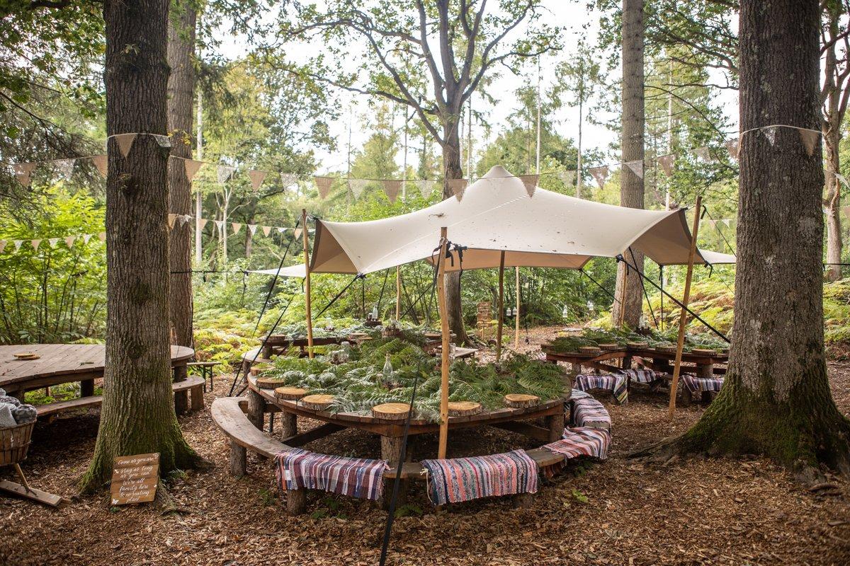 A canopy gazebo in the middle of the woods suspended over two round oak tables with benches either side dressed for a wedding with foliage, surrounded by bunting