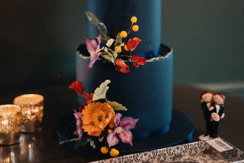 a three tier navy wedding cake with gold edging is decorated with bright flowers and has two grooms made out of icing next to it