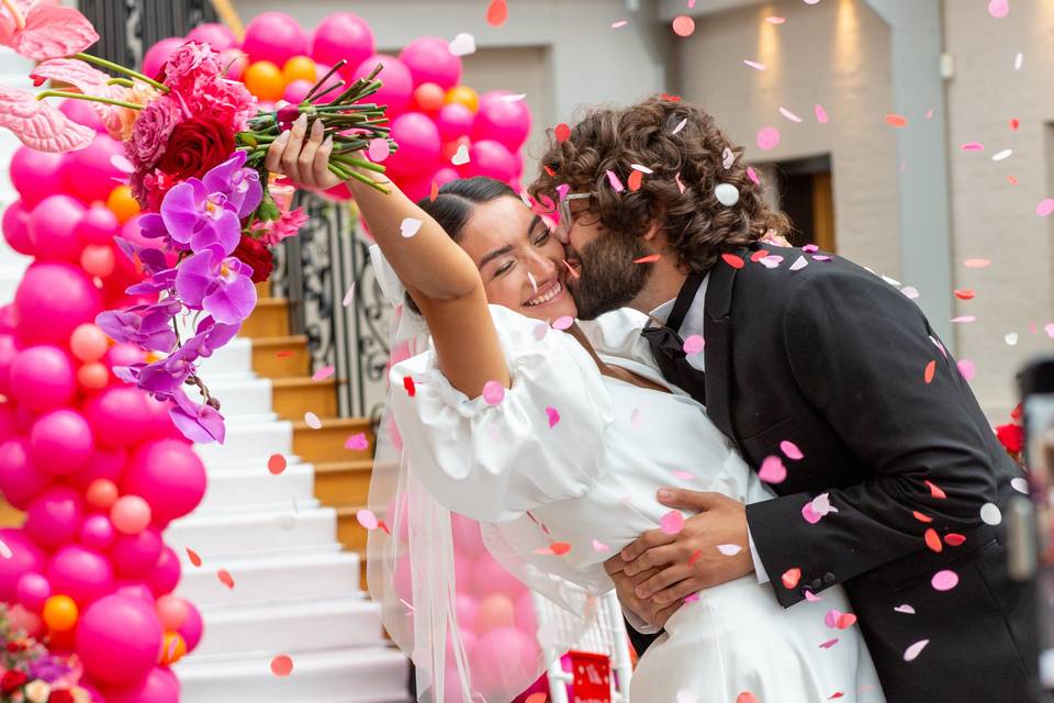 a groom dipping his wife as he kisses her on the cheek, as she dips back smiling and throwing her bouquet beneath a balloon arch