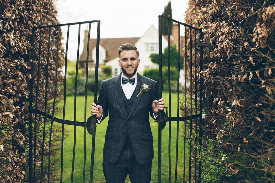 A groom in a grey check tuxedo suit leaning against a black railed fence in front of a countryside wedding venue