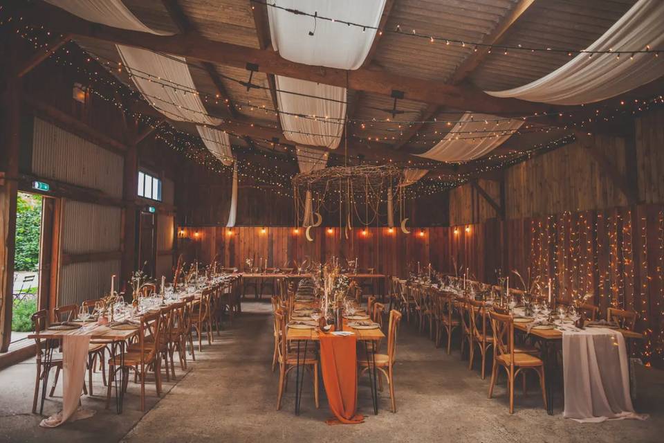 a rustic yorkshire wedding barn with three long banquet tables decorated beautifully and drapes and fairy lights hanging from the ceiling
