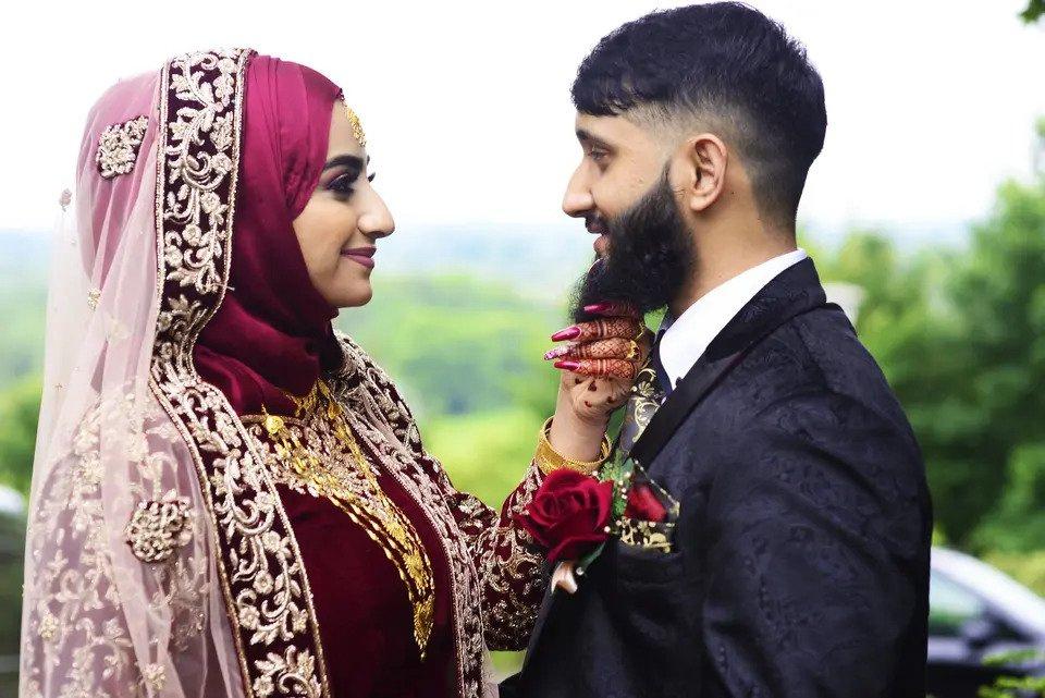 Bride wearing traditional Asian wedding attire place her hand under the chin of the suited groom opposite her with the countryside in the background