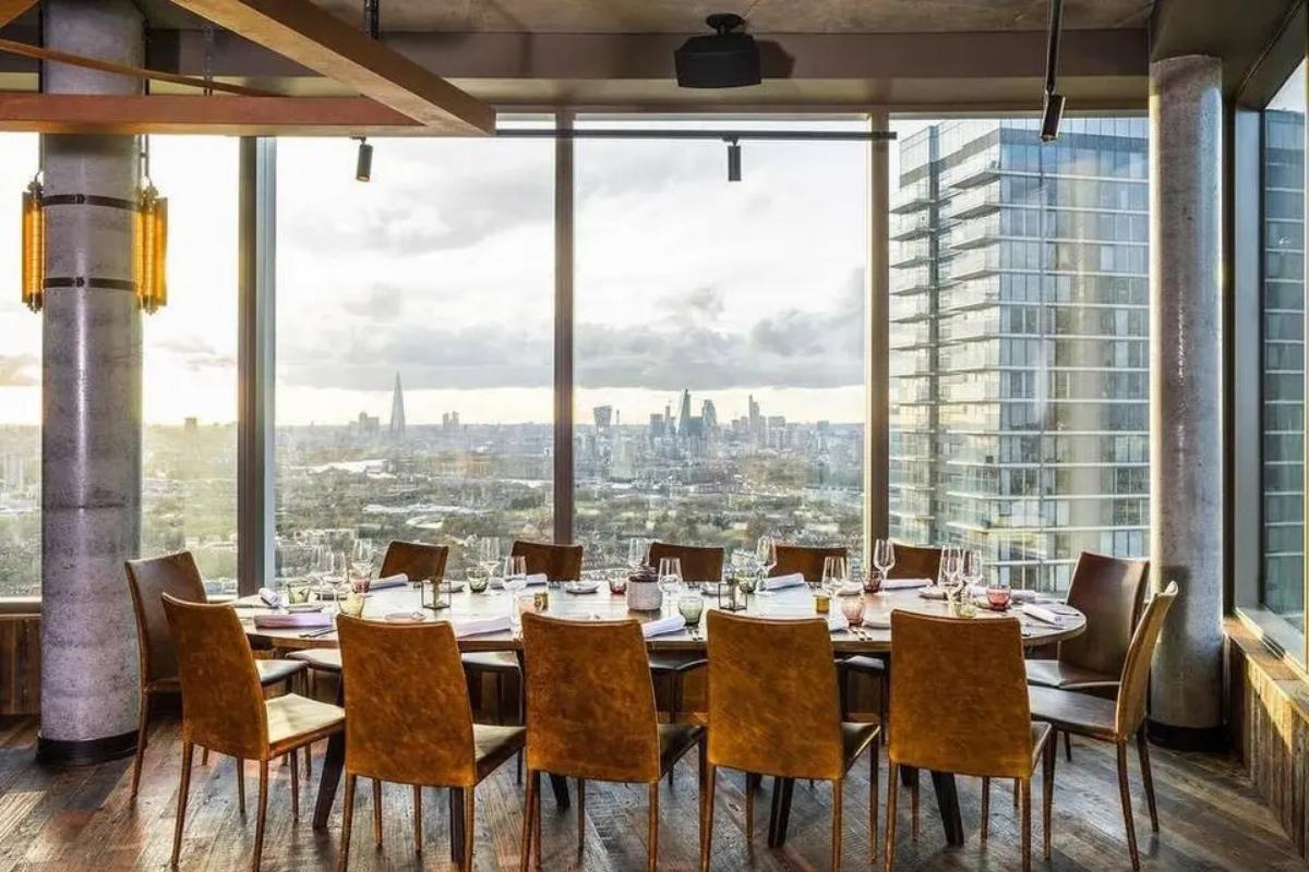  an oval table set up for 14 people with floor to ceiling windows showcasing the london skyline behind it