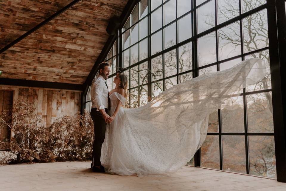 A bride and groom holding hands in front of a large, glass wall in an oak cabin, in front of a view of autumnal bare trees