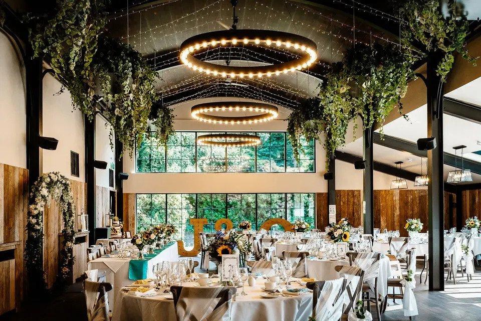 Barn dining room set for wedding meal with white round tables, Chiavari chairs, hanging chandeliers, hanging plants and floor-to-ceiling windows