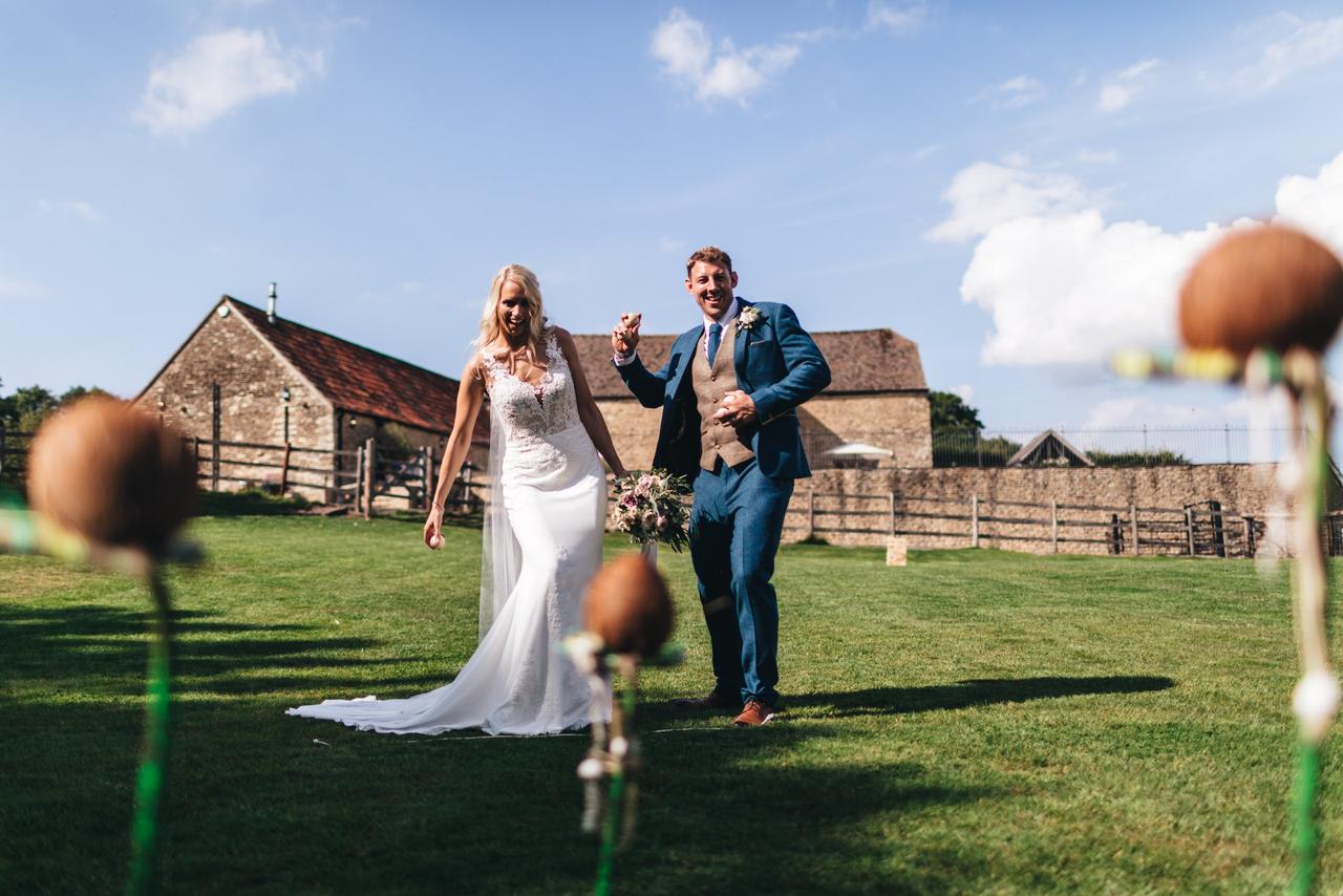 A bride and groom throwing balls at a coconut shy outside of a rustic barn wedding venue