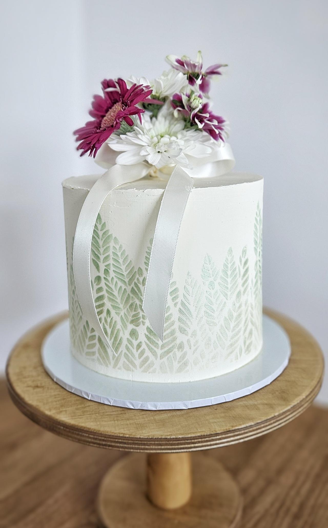 A white single tier wedding cake with subtle green leaf print and flowers on the top.