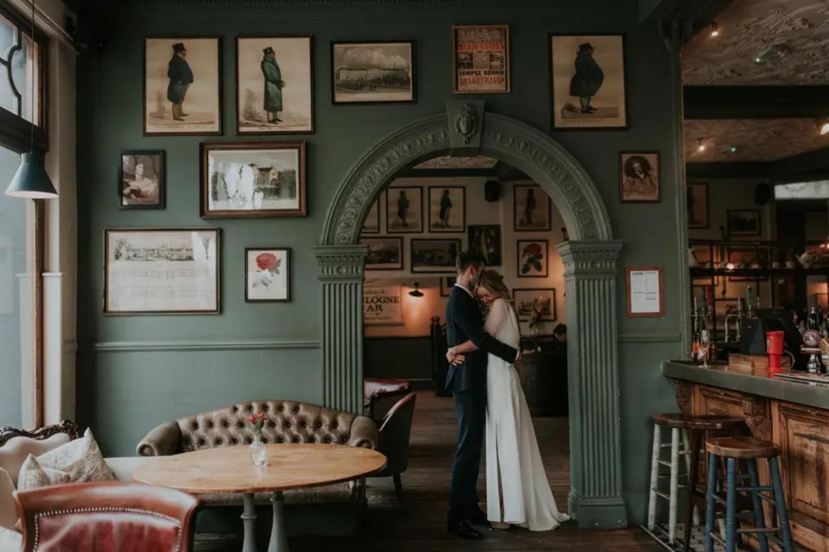  a couple hugging inside a small london pub wedding venue underneath a sage green arch 