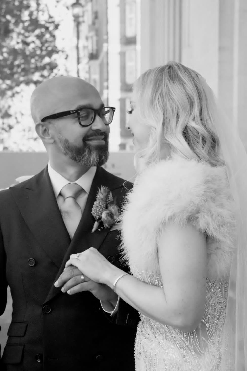 Black and white photo of a brown skinned groom with black glasses and a dark suit smiling at his blonde haired white bride who's wearing a cream fur stole and beaded ivory wedding dress