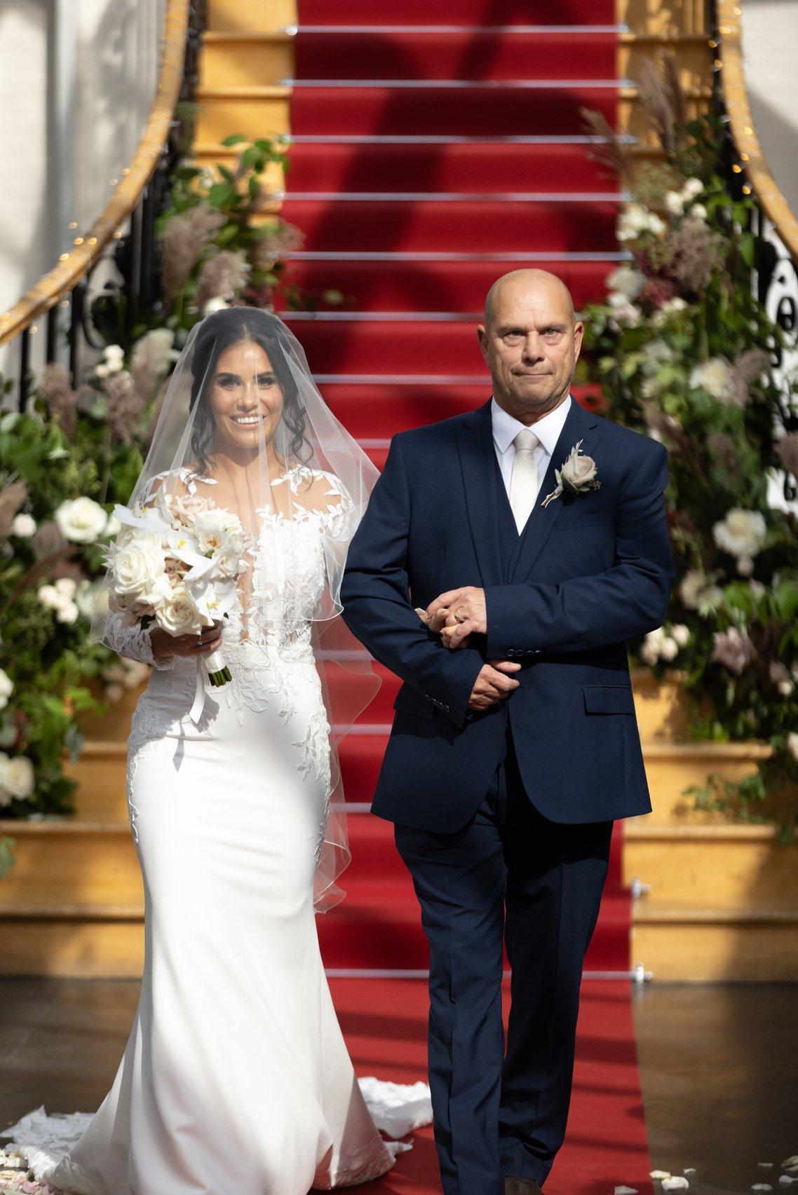 a bride links arms with her dad as they walk down the aisle with a red carpet and staircase behind them