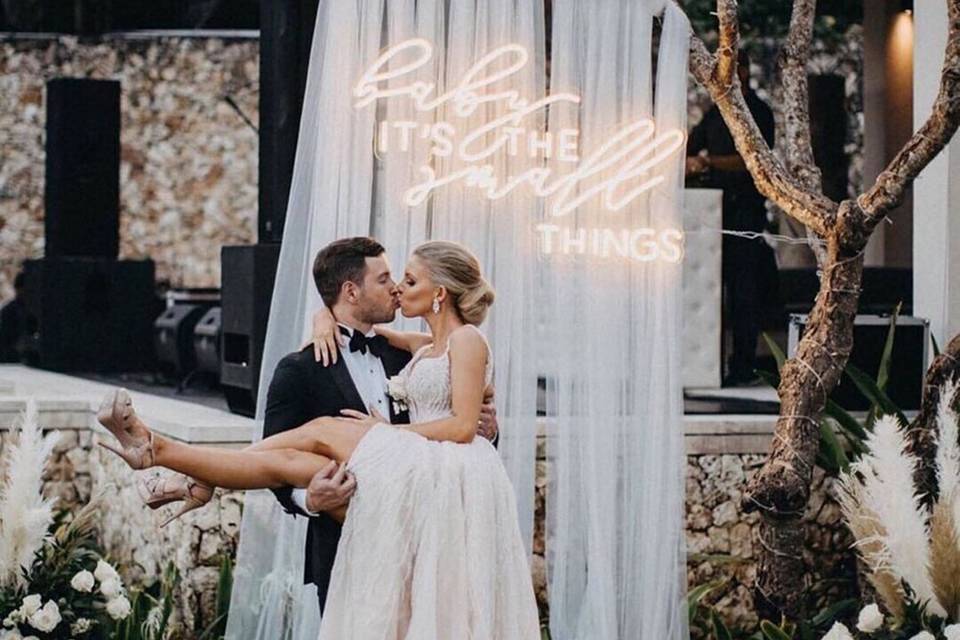A couple kisses in front of a photo backdrop, with white drapery, lots of flowers, and a neon sign