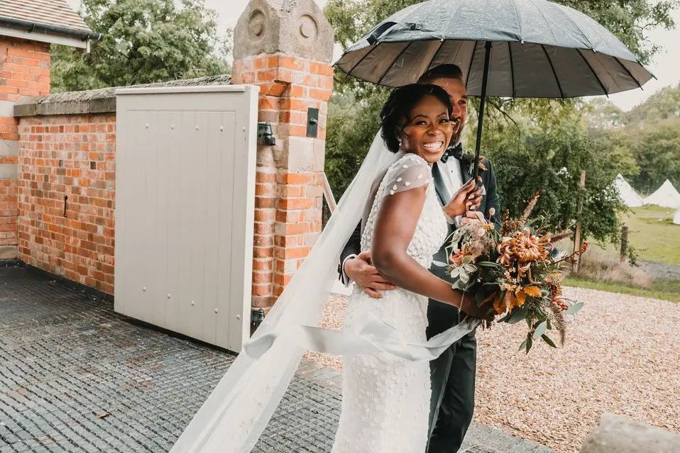 A bride is beaming at the camera as she looks over her shoulder towards it, whilst she and her groom are walking with an umbrella through the grounds of their wedding venue