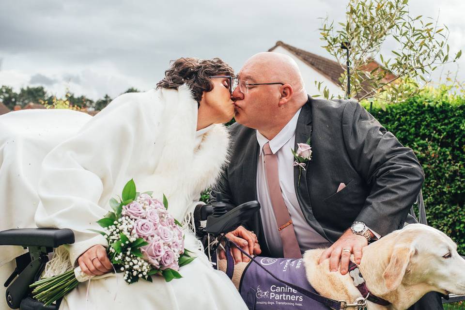 A bride in a wheelchair wearing a fur gilet and holding a lilac bouquet as she kisses a groom in a grey suit and red tie