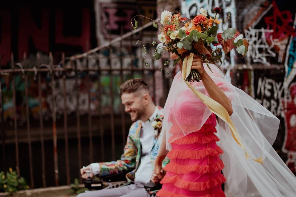 Newlywed couple celebrating their wedding - the groom is next to the bride in a motorised wheelchair, and the bride is holding the bouquet up by her face