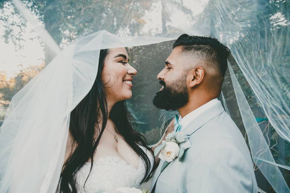Indian couple in a white wedding dress and light blue wedding suit smiling at each other under a sheer bridal veil