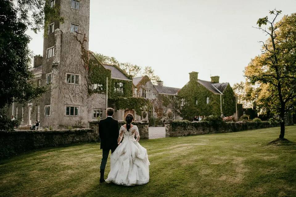 A couple is walking hand-in-hand towards an old ivy-covered stone castle, with manicured lawns and old trees around it.