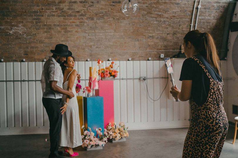 A wedding content creator captures pictures of a bride and groom posing next to a wedding cake on her mobile phone