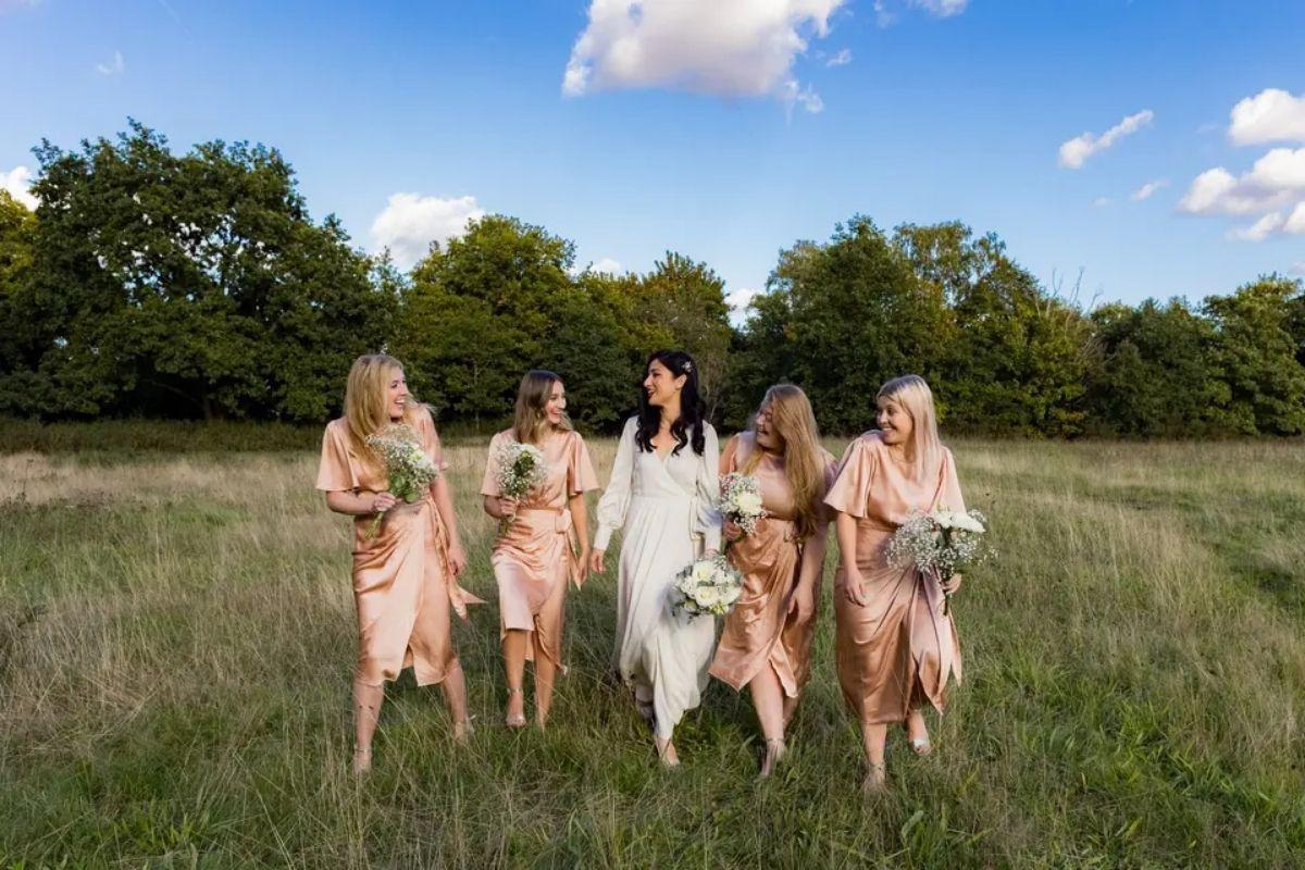 a bride and her four bridesmaids in peach bridesmaid dresses all hold their bouquets and walk through a field