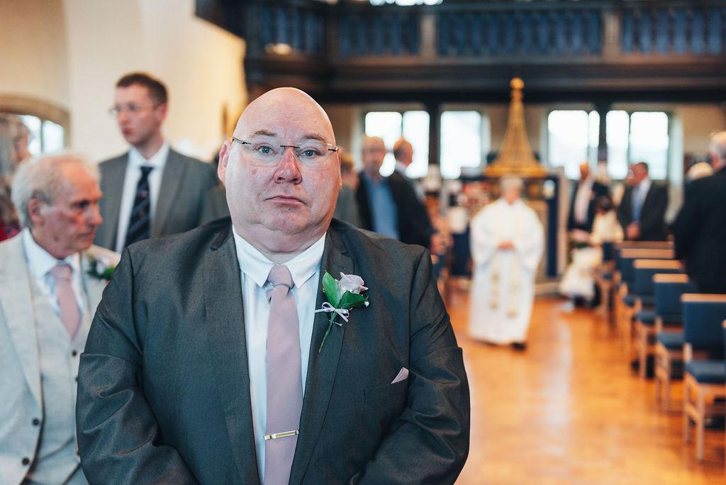 A groom standing at the altar with his hands held in front of him and guests behind him