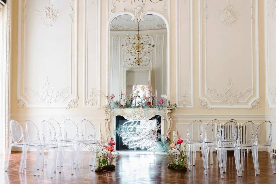 A lavish wedding ceremony set up with ornately decorated walls and clear chairs in front of an original fireplace, decorated with pink florals and a neon sign reading 'happily ever after'