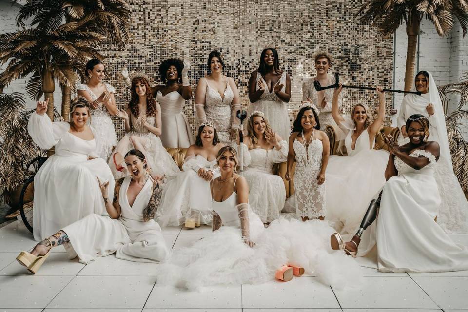 Group shot from a diverse wedding shoot featuring a range of different models in wedding dresses, including a heavily tattooed woman, a model in a wheelchair, a trans model, a model with one leg, a model with one arm, a model with Downs Syndrome, a model holding her walking stick up in the air and more, all posed in front of gold palm trees and a glittering gold back drop in an industrial wedding venue setting