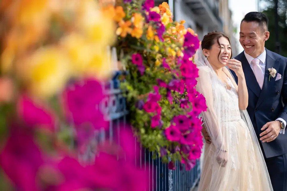 Bride and groom laugh in front of a wrought iron fence with flowers