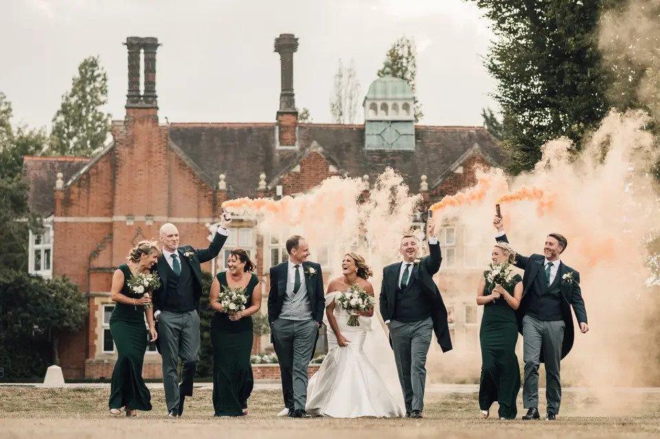 Bride and groom walking away from the venue with their bridesmaids and groomsmen as the groomsmen hold up peach coloured smoke cannons