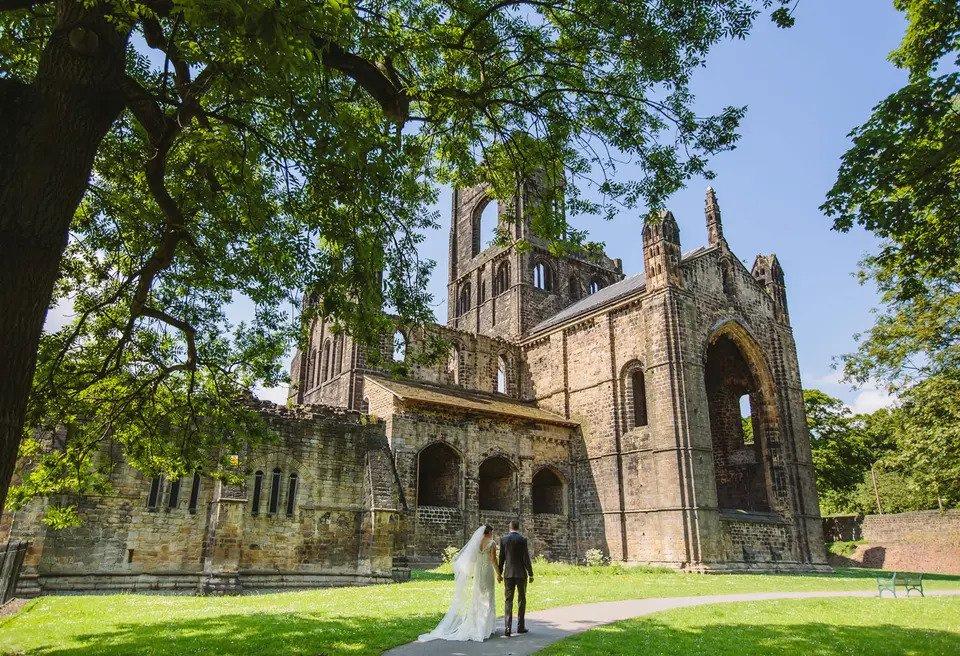  A couple walk down a path across a lawn towards the gothic Kirkstall Abbey