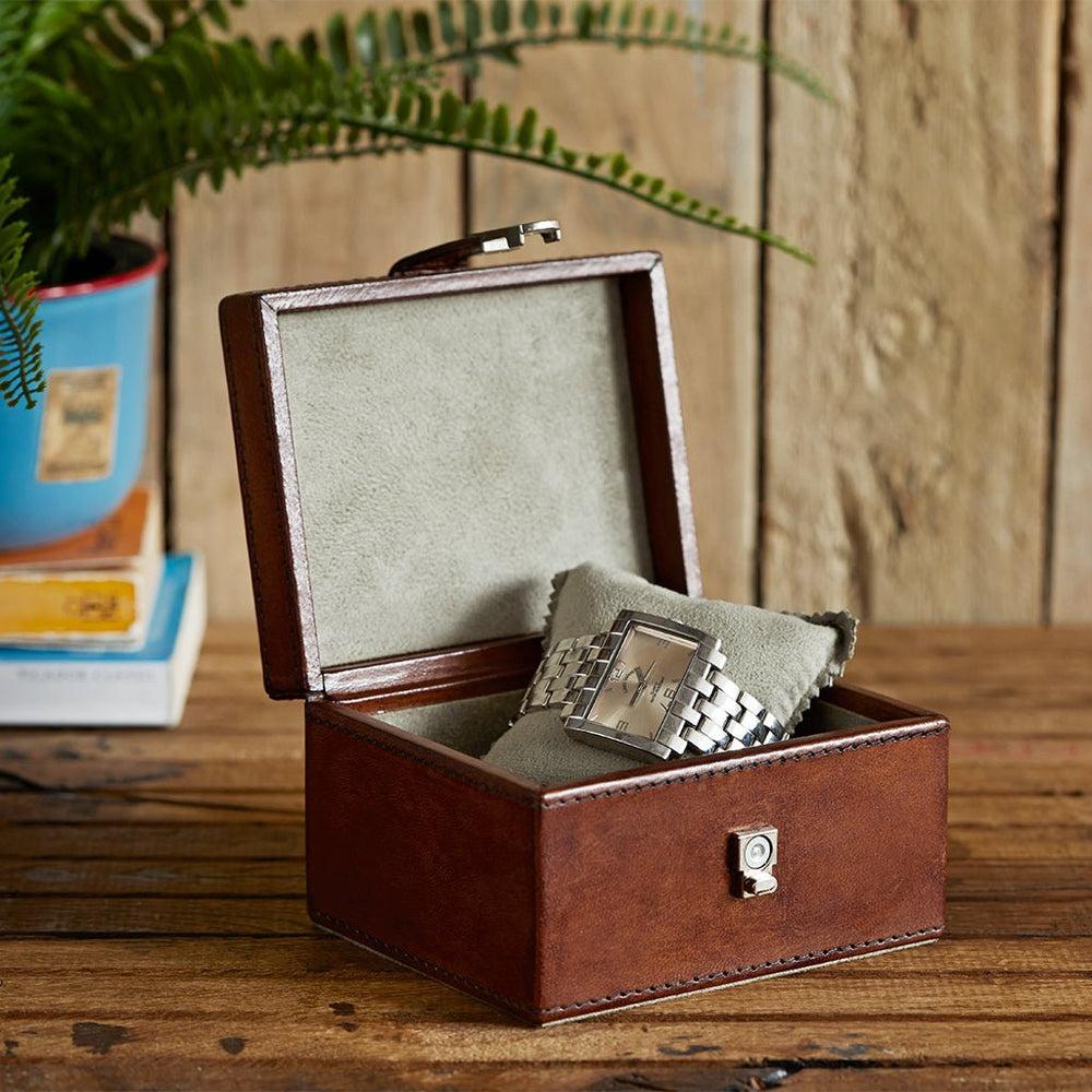 Image of a brown leather watch box with chrome clasp on a table. It is open with a watch inside.