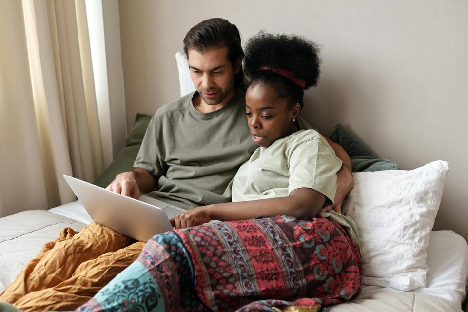 A couple sitting on a bed together making a wedding website on their laptop