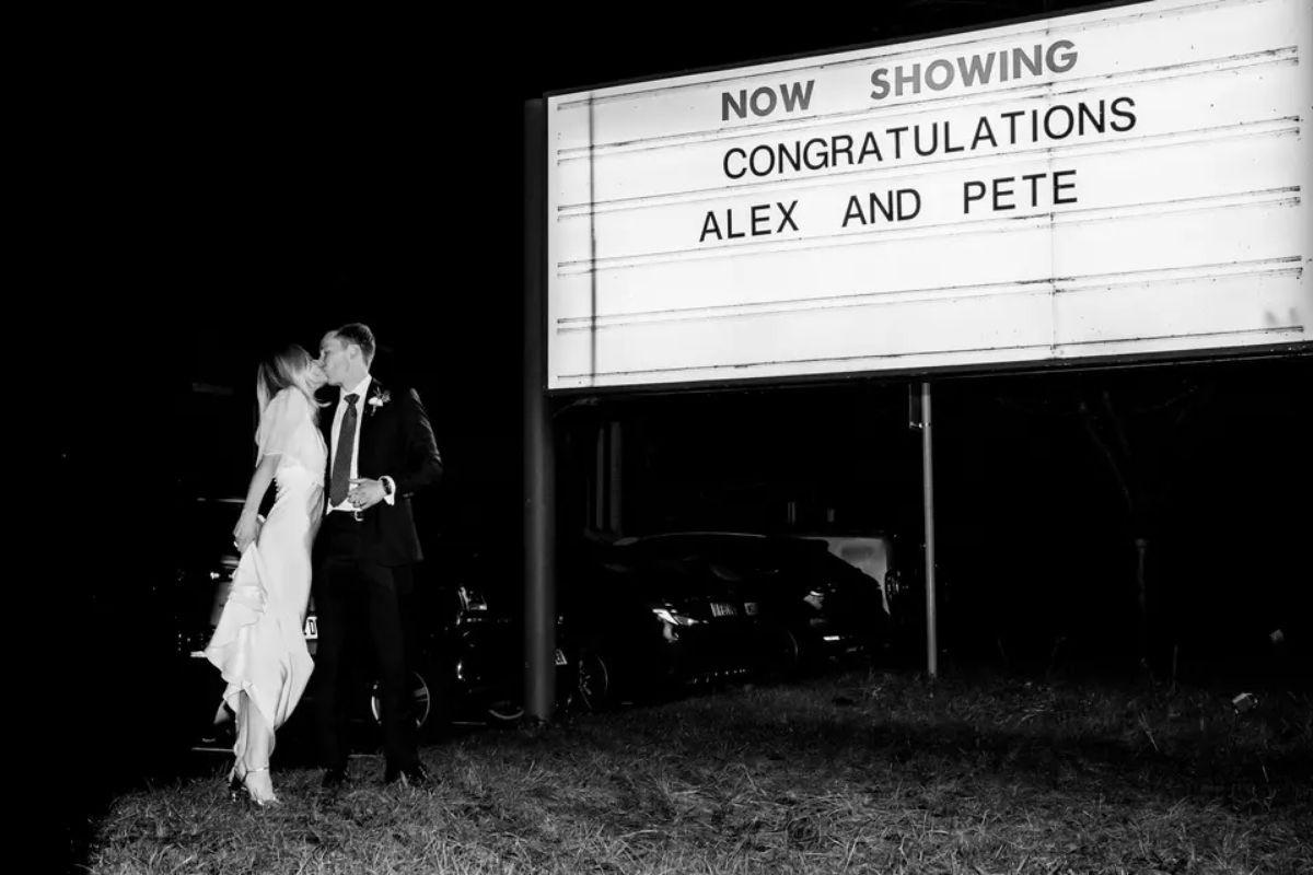 a bride and groom pose for a black and white picture next to an old cinema sign that says 'Now showing - congratulations - alex and pete'