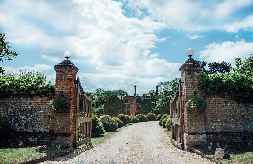 A brick wall and wooden gate opens up onto a long walkway which leads up to a grand wedding venue