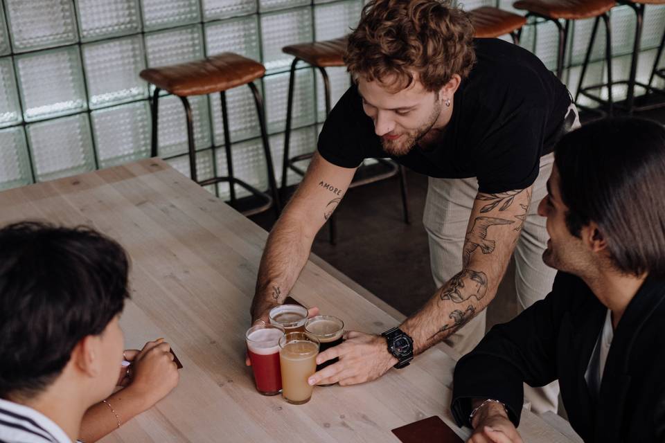 Four men sat at a pale oak table in a cafe with one bringing over four alcoholic drinks about to plan a stag do