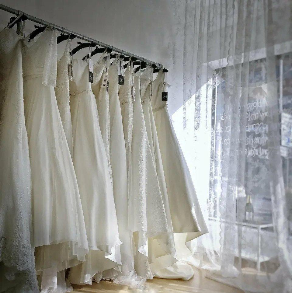 Wedding dresses hanging in a shop in front of a lace curtain and window