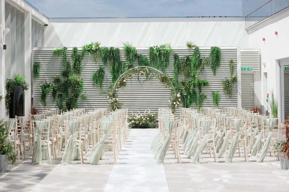 Wedding ceremony set up in a patio with wooden chairs with green sashes, plants and a floral hoop