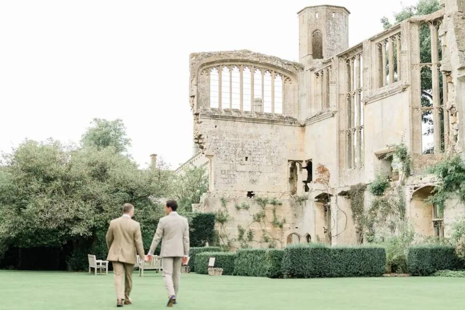two grooms holding hands walking along the lawns next to a light stone luxury castle wedding venue