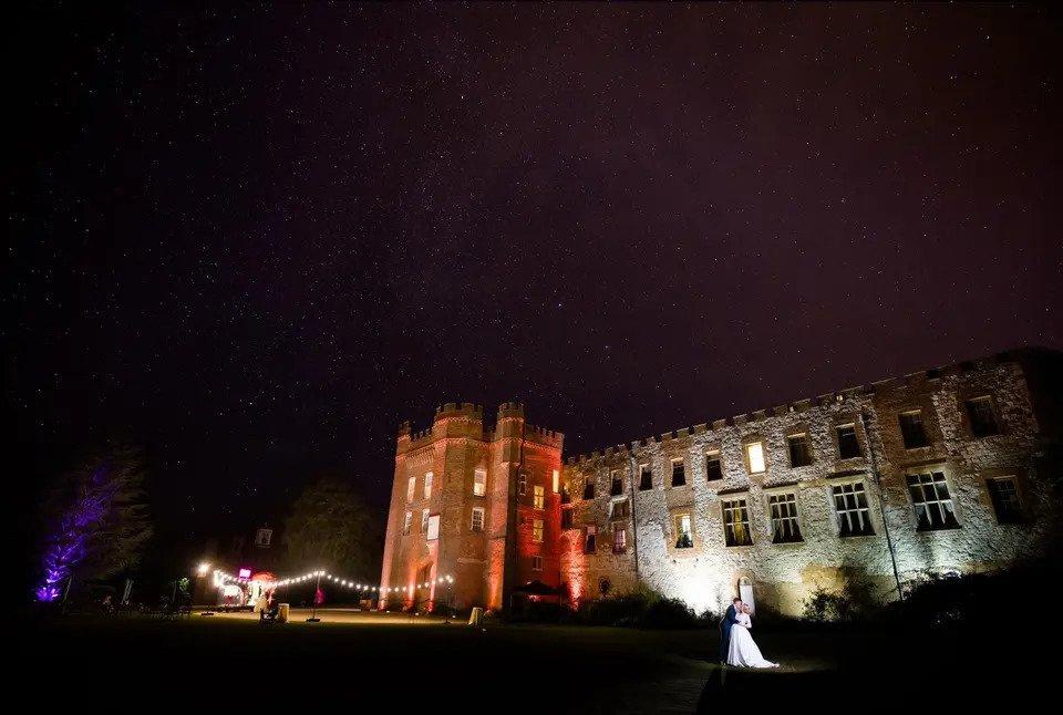 A night-time image of Farnham Castle lit up with fairy lights and featuring a bride and groom