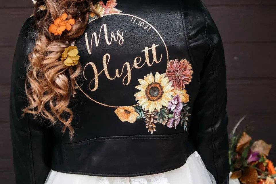 The back of a bride wearing a black leather jacket saying 'Mrs Higell'. Her long hair is in a loose fairytale braid decorated with colourful flowers