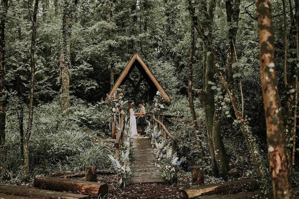 A rustic wedding venue with a wooden bower in the forest where a bride and groom are exchanging vows