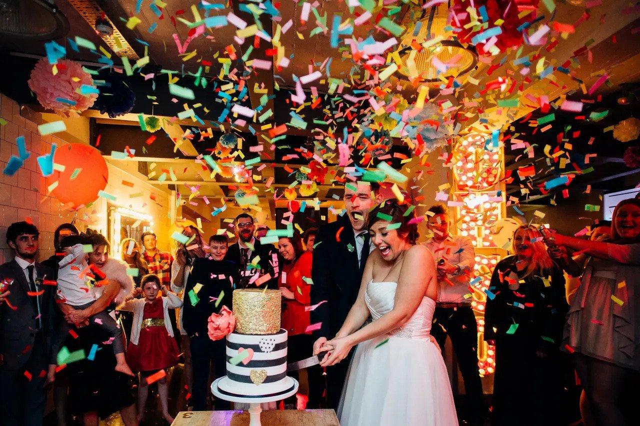 Bride and groom cutting into a wedding cake as confetti is tossed over them